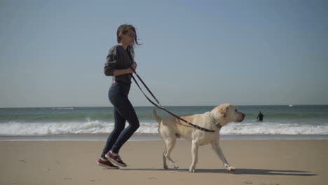 Smiling-woman-in-eyeglasses-running-with-labrador-on-beach.