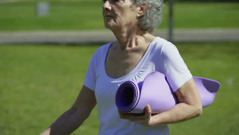 Side-view-of-woman-walking-in-park,-holding-yoga-mat-in-hand