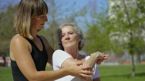 Focused-elderly-lady-with-coach-stretching-arm-in-park-.