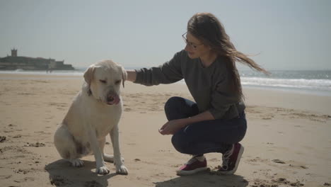 Smiling-beautiful-lady-in-eyeglasses-sitting-on-sandy-beach-with-pet.