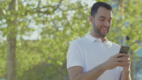 Smiling-young-man-strolling-in-park-with-smartphone-in-hands.