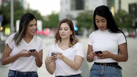 Joyful-friends-walking-on-street-and-texting-on-smartphones