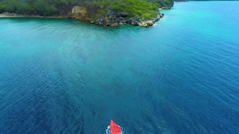 Drone-follows-boat-and-wake-in-open-ocean,-tilt-up-to-rocky-coastline-of-curacao