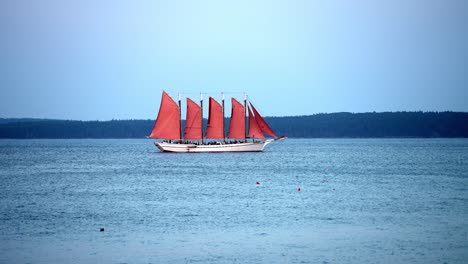 Large-red-multi-mast-sailboat-cruises-along-blue-lake-with-overcast-skies,-slow-motion