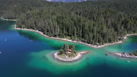 Fly-over-little-small-tiny-island-in-the-middle-of-lake-and-yacht-club-boating-in-crystal-clear-turquoise-blue-green-shallow-water-in-Eibsee-bayern-Deutschland-pine-green-tree-forest-around-the-lake