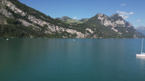 Eine-Weiße-Bootsjacht-Schwimmt-In-Einer-Ruhigen-Blauen-See--Und-Berglandschaft-In-Den-Alpen,-Einem-Schweizer-Alpendorf-In-Der-Schweiz,-Das-über-Den-Malerischen-Horizont-Fliegt.-Drohnenaufnahme-In-Geringer-Höhe-In-Der-Touristischen-Sommersaison-In-Europa