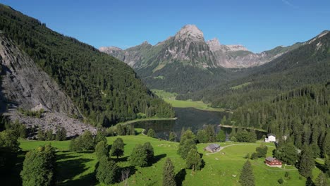 Scenic-nature-landscape-of-Swiss-wooden-cabin-chalet-farm-on-green-alpine-meadow-surrounded-by-alp-background-mountains-pine-trees-overlook-turquoise-glacier-lake-in-a-sunny-day-in-Europe-Switzerland