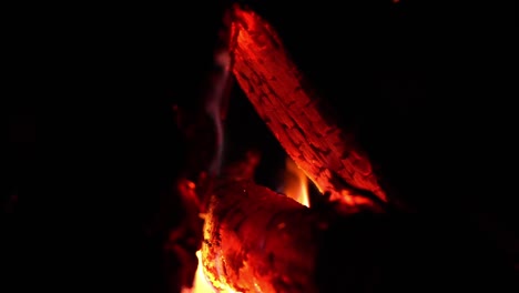 Side-view-into-glowing-fire-with-flames-rising-around-burning-log,-smoke-releases-into-darkness