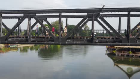 Drone-sliding-to-the-right-revealing-people-crossing-the-River-Kwai-Memorial-Bridge-curiously-looking-around-this-World-War-II-museum-of-a-structure,-Kanchanaburi,-Thailand
