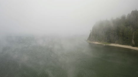 Aerial-view-over-misty-water,-passing-the-lakeshore-and-a-beach-at-a-lake,-in-USA