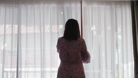 A-woman-talking-on-her-mobile-phone-is-seen-from-her-back-almost-as-a-silhouette-at-the-window-in-a-room-as-she-reaches-out-for-the-stick-to-adjust-the-curtain