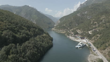 Disparo-De-Un-Dron-Volando-Sobre-Las-Montañas-Cerca-Del-Lago-Koman-En-Albania-En-Un-Día-Soleado-Con-Nubes-Con-Agua-Azul-Y-Un-Valle-Verde-Y-Un-Muelle-De-Ferry-En-El-Registro-De-Vistas