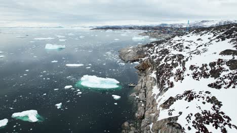 Rocky-coast-with-flowing-icebergs-and-town-of-Ilulissat-in-distance,-aerial-view