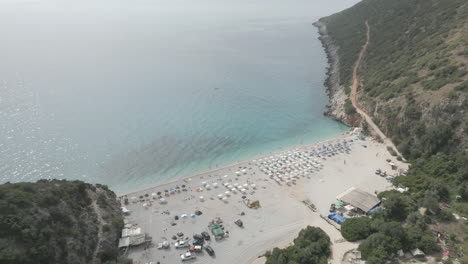 Drone-shot-over-Gjipe-beach-Albania-with-tourists-on-the-beach-and-clear-blue-water-near-the-mountains-and-the-sea-on-a-sunny-and-bright-day-and-green-nature-around-with-sun-beds-towels-LOG
