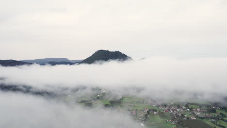Luftdrohnenflug-Durch-Wolken-Mit-Einem-Berggipfel-Im-Hintergrund-Und-Einem-Kleinen-Dorf-Darunter