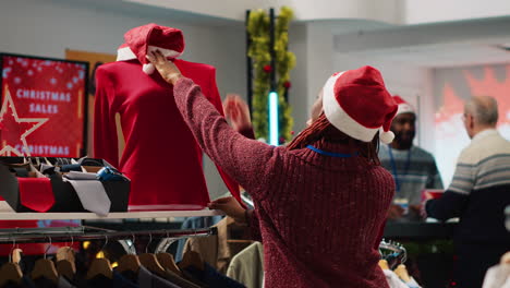 African-american-retail-assistant-wearing-Santa-hat-helping-asian-client-browsing-through-festive-clothing-in-Christmas-shopping-store-during-winter-holiday-season.-Employee-assisting-customer