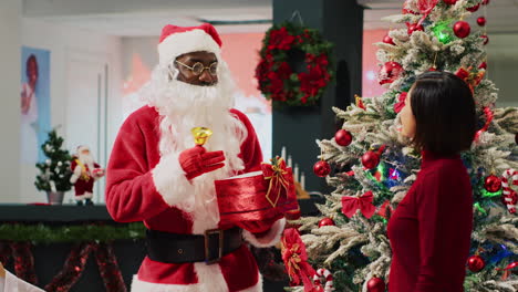 African-american-employee-dressed-as-Santa-Claus-ringing-bell-and-holding-xmas-gift-next-to-xmas-ornate-tree,-talking-with-asian-client-shopping-in-festive-decorated-fashion-store