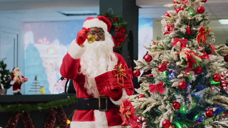 African-american-employee-posing-as-Santa-Claus-ringing-bells-next-to-beautifully-decorated-Christmas-tree,-trying-to-attract-customers-to-fashion-store-during-promotional-holiday-season