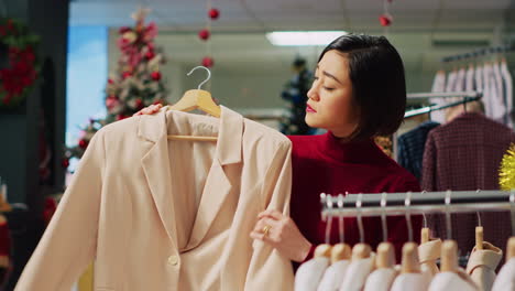 Portrait-of-happy-woman-browsing-through-elegant-blazers-on-racks-in-Christmas-adorn-clothing-store-during-winter-holiday-season.-Smiling-client-checking-fashion-boutique-items