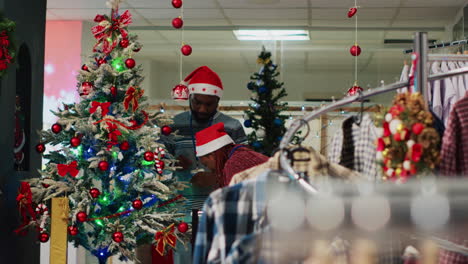 African-american-employees-wearing-Santa-hat-ornating-Christmas-tree-in-clothing-store-before-festive-promotional-event.-Retail-assitants-ornating-fashion-shop-during-winter-holiday-season