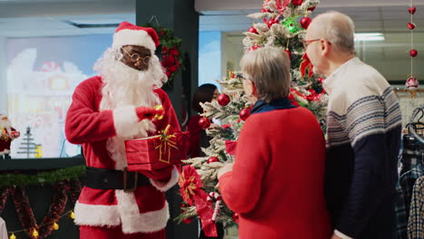 African-american-worker-dressed-as-Santa-Claus-ringing-bell-and-holding-xmas-gift-next-to-beautifully-decorated-Christmas-tree,-talking-with-senior-couple-shopping-in-xmas-decorated-fashion-store