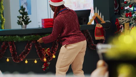 African-american-employee-wearing-Santa-hat-decorating-clothing-store-with-Christmas-ornaments-before-festive-promotional-event.-Worker-installing-xmas-tree-in-fashion-shop,-helped-by-colleague