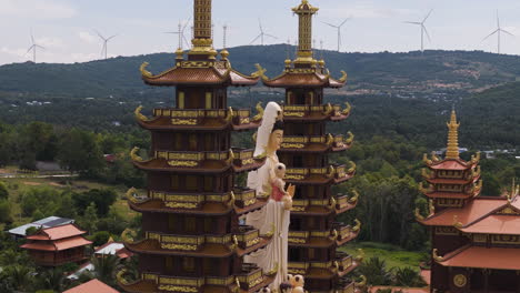 Orbital-shot-of-Buddhist-temple-with-two-pagoda-towers-and-wind-turbines,-aerial-closeup