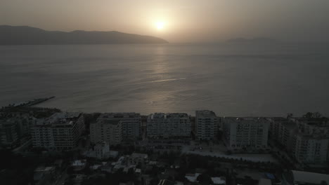Drone-shot-above-the-city-Vlore-Albania-looking-over-the-buildings-and-harbour-with-ferry-with-the-sea-and-beach-underneath-during-sunset-in-the-evening-with-an-orange-glow-LOG