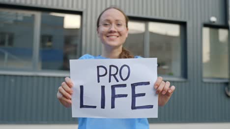 Professional-Female-Worker-with-a-Clear-Statement-Holding-Paper-Reading-"Pro-Life"-in-Front-of-a-Building-Outside-with-Close-Up-Shot