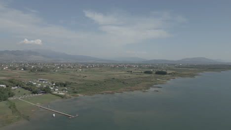 Drone-shot-flying-over-Shkodra-Lake-in-Albania-with-mountains-in-the-background-and-green-plants-grass-nature-underneath-on-a-sunny-day-with-haze-above-the-lake-LOG