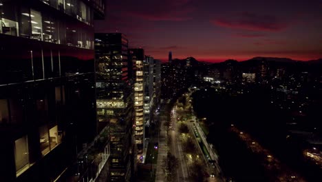Überflug-über-Die-Modernen-Gebäude-Von-Nueva-Las-Condes-In-Santiago,-Chile,-Dem-Geschäftszentrum-Der-Stadt,-Mit-Dem-Costanera-Turm-Im-Hintergrund-Bei-Nacht-Und-Einem-Orangefarbenen-Sonnenuntergang