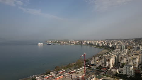 Drone-shot-hyperlapse-above-the-city-Vlore-Albania-looking-over-the-buildings-and-harbour-with-ferry-with-the-sea-and-beach-underneath-on-a-sunny-day-with-a-little-bit-of-haze-LOG
