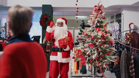 African-american-man-dressed-as-Santa-Claus-in-Christmas-themed-shopping-mall-clothing-store.-Employee-jingling-xmas-bells-and-holding-red-package-present-in-festive-decorated-shop