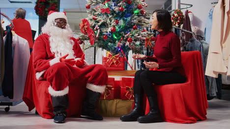 African-american-employee-acting-as-Santa-Claus-sitting-down-next-to-Christmas-tree-with-asian-woman-in-xmas-ornate-clothing-store.-Fashion-shop-worker-offering-gift-to-customer