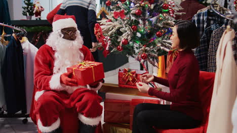 African-american-manager-acting-as-Santa-Claus-sitting-down-next-to-Xmas-tree-with-woman-in-festive-decorated-clothing-store.-Fashion-boutique-supervisor-offering-present-to-grateful-client