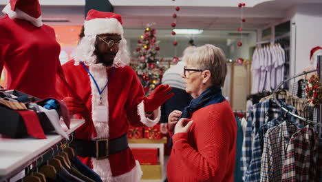 Manager-wearing-Santa-Claus-costume-around-shopping-mall-fashion-boutique-chatting-with-elderly-woman.-Supervisor-in-holiday-themed-suit-showing-senior-client-red-garment-piece