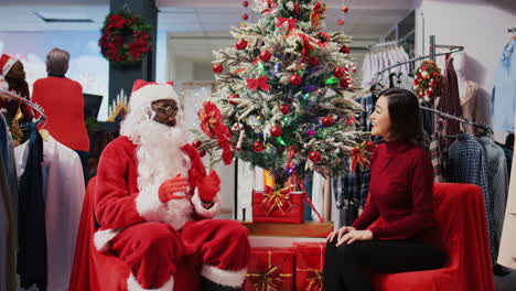 Dolly-out-shot-of-worker-acting-as-Santa-Claus-sitting-down-next-to-Christmas-tree-with-asian-woman-in-xmas-decorated-clothing-store.-Fashion-shop-retail-assistant-offering-present-to-customer