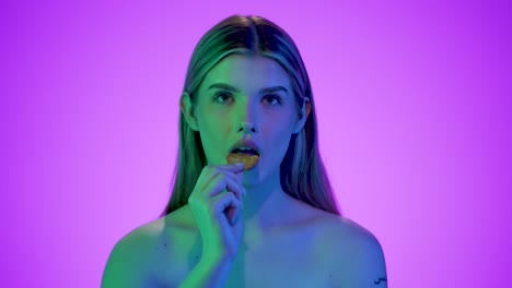 Medium-shot-of-pretty-beautiful-woman-holding-a-fluted-potato-chip-into-the-camera-and-then-eating-it-with-relish-in-front-of-a-purple-background-with-blue-contrasts-in-her-face-in-slow-motion