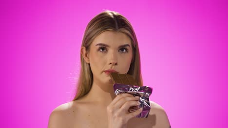 Medium-shot-of-a-beautiful-young-woman-or-model-while-she-enjoys-a-bite-of-the-vegan-chocolate-bar-and-loves-different-candy-sweets-against-purple-background-in-slow-motion