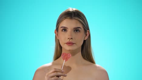 Medium-Static-slow-motion-shot-of-a-young-caucasian-naked-woman-licking-a-heart-shaped-lollipop-while-looking-sexy-into-the-camera-in-slow-motion-against-turquoise-background