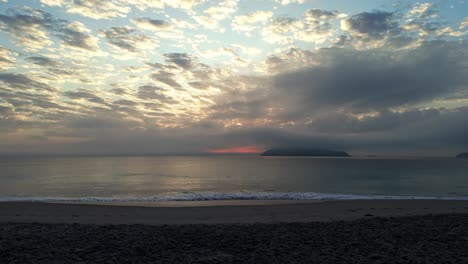 a-beach-with-a-few-clouds-and-a-mountain-in-the-distance-with-a-sun-rising-in-the-distance-behind-it