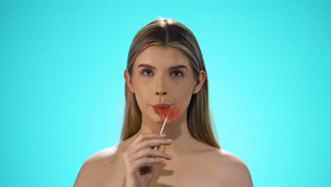 Static-shot-of-a-young-caucasian-naked-woman-licking-a-heart-shaped-lollipop-while-looking-sexy-into-the-camera-in-slow-motion-against-turquoise-background