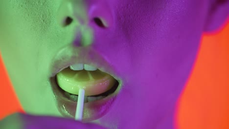 Close-up-shot-of-young-sexy-woman-lips-while-she-licks-a-sweet-lollipop-with-pleasure-with-her-tongue-against-orange-background-with-green-purple-contrast-on-face-in-slow-motion