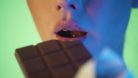Close-up-shot-of-beautiful-young-woman-lips-enjoying-and-lewdly-taking-a-bite-of-a-delicious-chocolate-against-green-background-with-blue-contrast-in-her-face-in-slow-motion