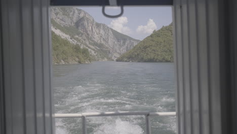 Eine-Fährfahrt-In-Albanien-Von-Fierze-Nach-Koman-über-Den-See-Mit-Einer-Majestätischen-Natur-Rundherum-An-Einem-Sonnigen-Tag-Mit-Wolken-Und-Blick-Auf-Den-Fluss,-Gesehen-Von-Der-Rückseite-Des-Bootes-Zwischen-Zwei-Baumstämmen