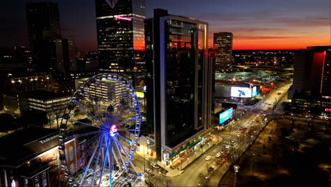 Aerial-view-of-ferris-wheel,-modern-skyscraper-and-Lightning-billboard-in-city-along-busy-intersection-of-Atlanta-during-dramatic-sunset