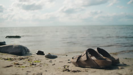 Women-picks-up-sandals-on-stone-beach-along-the-ocean-on-a-sunny-day,-Sandvik,-Öland-Sweden-,-close-up-wide