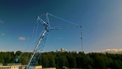 Full-view-of-a-blue-luffing-construction-crane-against-a-blue-sky-with-forest-in-background
