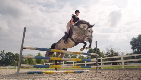 Girl-on-a-dapple-gray-horse-practicing-jumping-over-a-fence,-handheld-shot