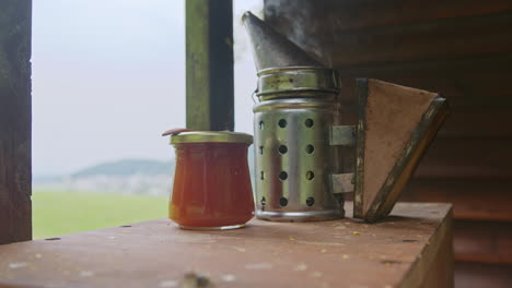 Bee-smoker-and-a-jar-of-sweet-honey-on-the-wooden-surface,-close-up-shot